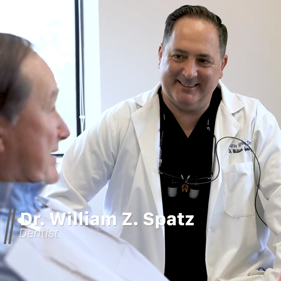 Dr. William Z. Spatz smiling while speaking with patient in dental office setting.